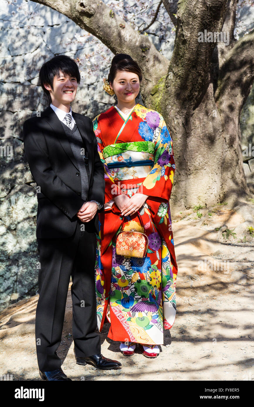 Giapponese giovane coppia sposata. Uomo in formale stile occidentale suit, woman in red kimono. In piedi sotto la fioritura dei ciliegi tree, sorridente. Nessun contatto visivo. Foto Stock
