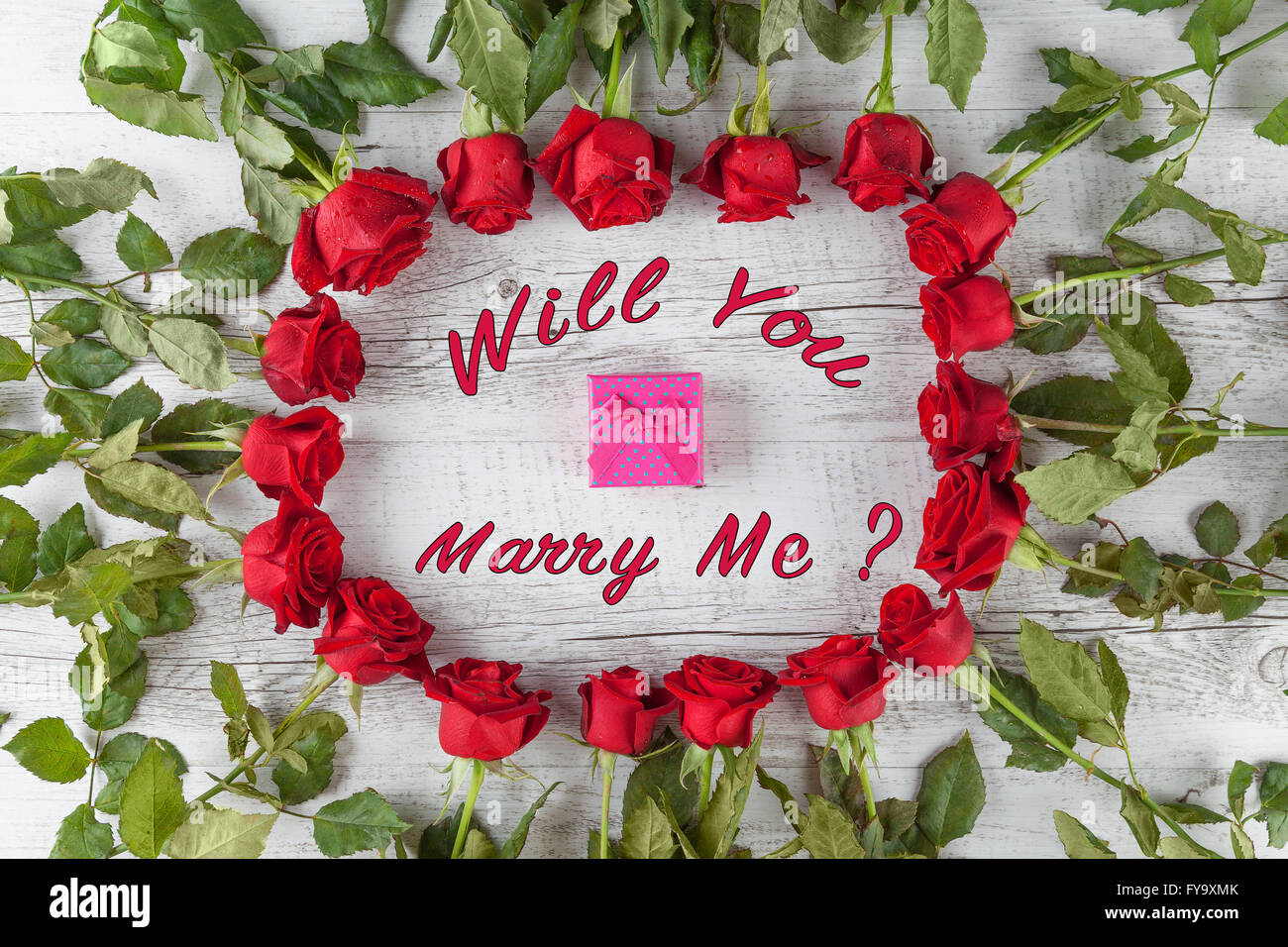 Ti sposerà me? Il testo con un piccolo regalo in un cerchio di rose rosse su bianco tavolo in legno. Vista dall'alto. Foto Stock