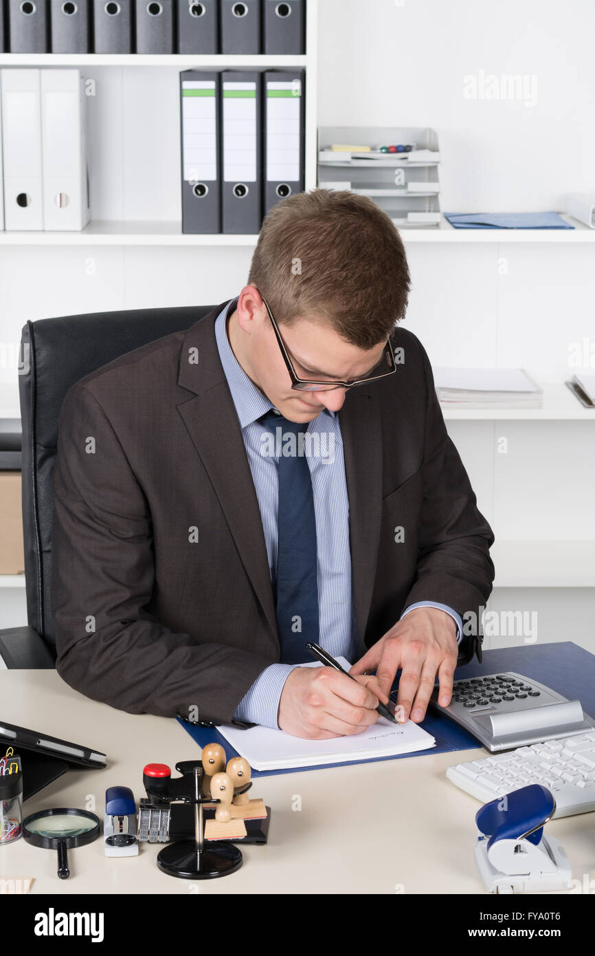 Giovane imprenditore è la scrittura in un file mentre seduto alla scrivania in ufficio. L'uomo è alla ricerca per il file. Foto Stock