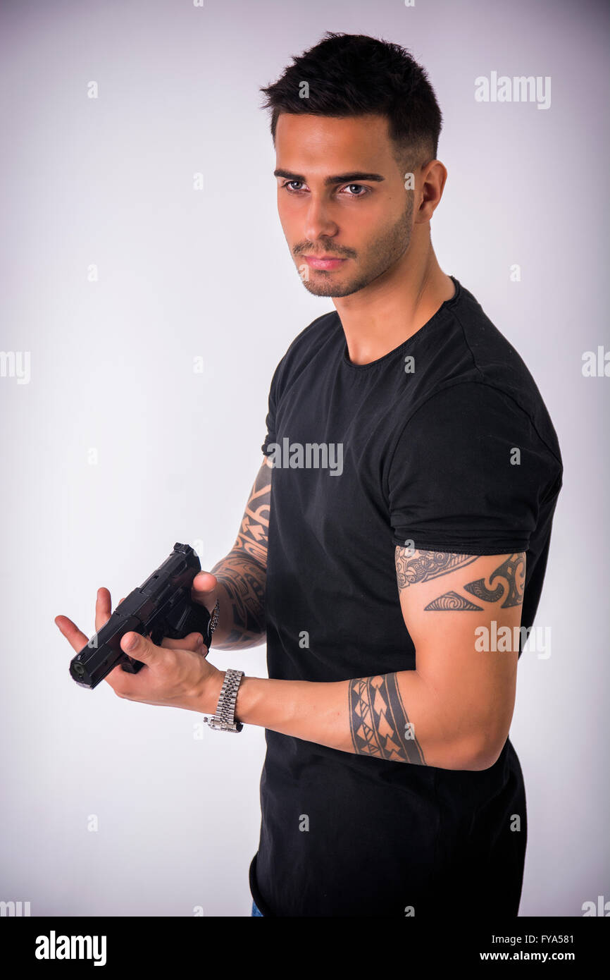 Giovane uomo bello puntando la pistola, indossando t-shirt nera su sfondo chiaro Foto Stock