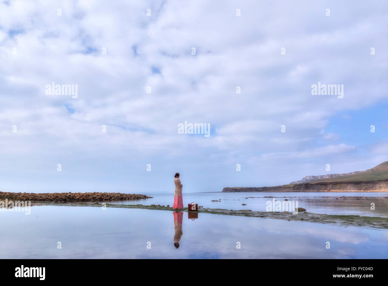 Una donna in un vintage abito rosa è in piedi al mare con la sua valigia Foto Stock