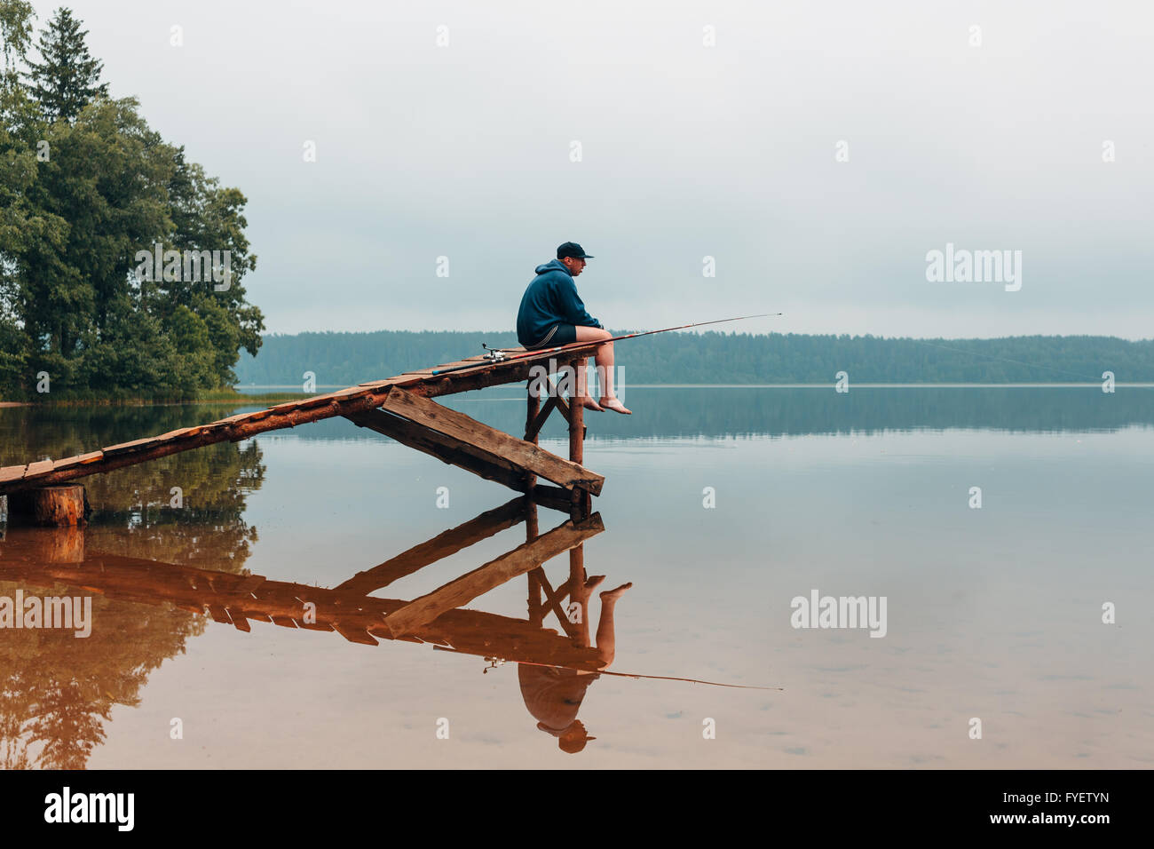 Uomo si siede su un ponte di legno attende quando i pesci sono di mordere. Foto Stock