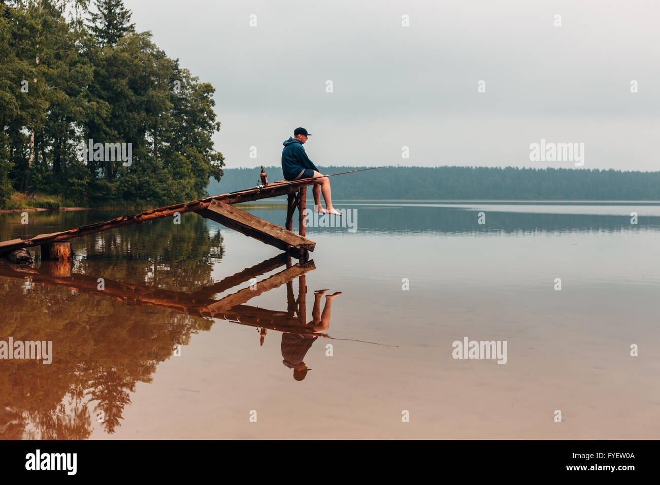 Uomo si siede su un ponte di legno attende quando i pesci sono di mordere. Foto Stock