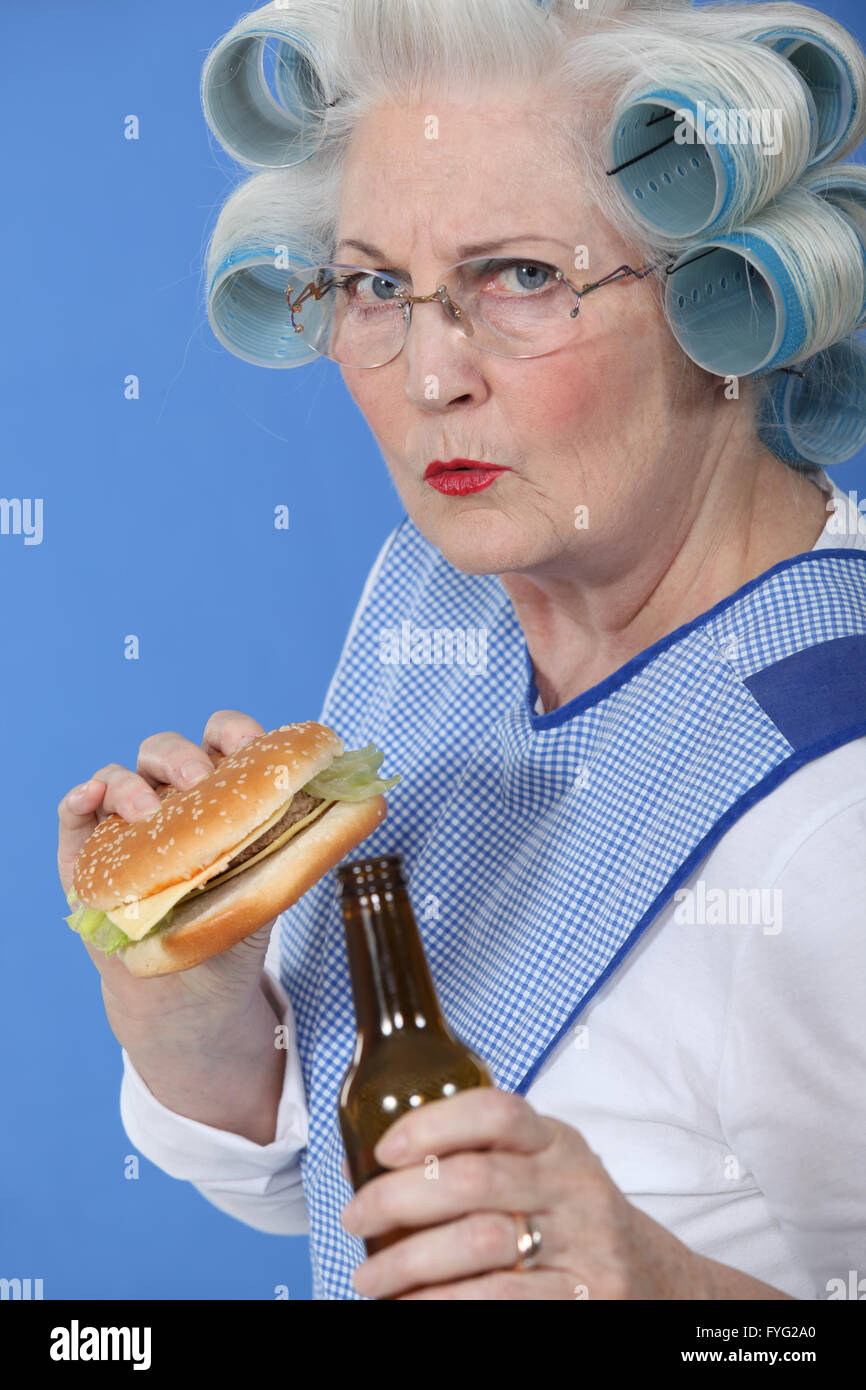 Senior con bigodini nei suoi capelli di bere birra e mangiare hamburger Foto Stock