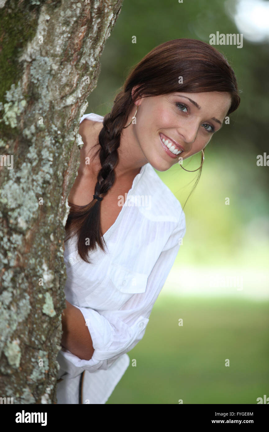 Donna sorridente sporgersi da dietro a un albero Foto Stock