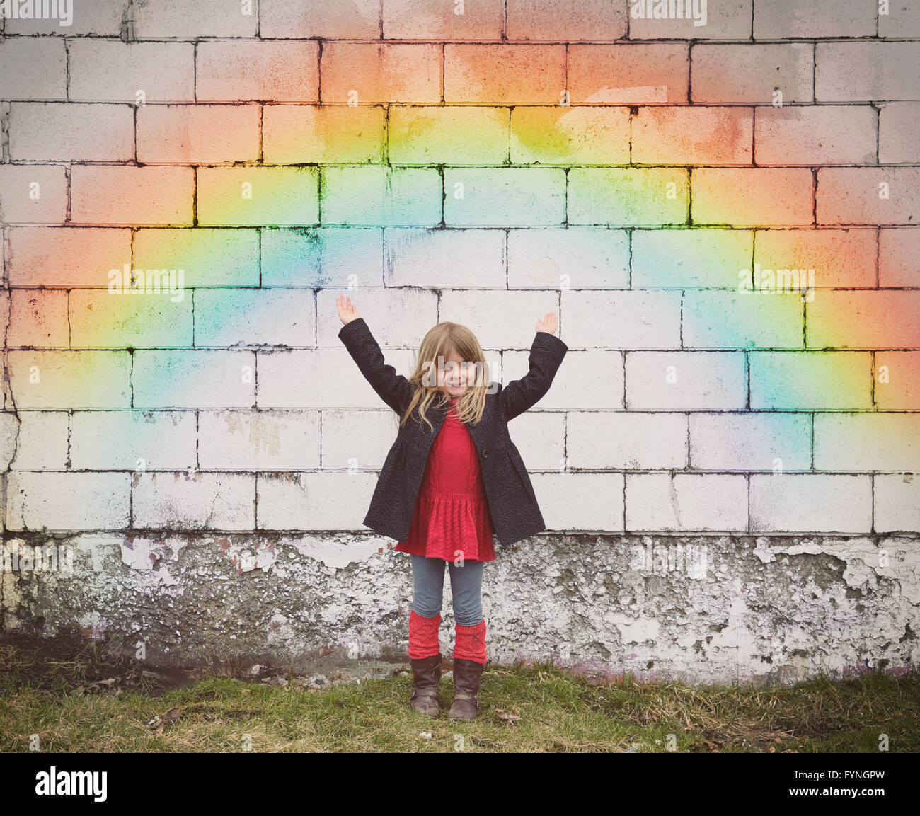 Un felice bambina è in piedi contro un vecchio muro di mattoni con un colorato arcobaleno e le sue mani sono sollevati per un concetto meteo Foto Stock