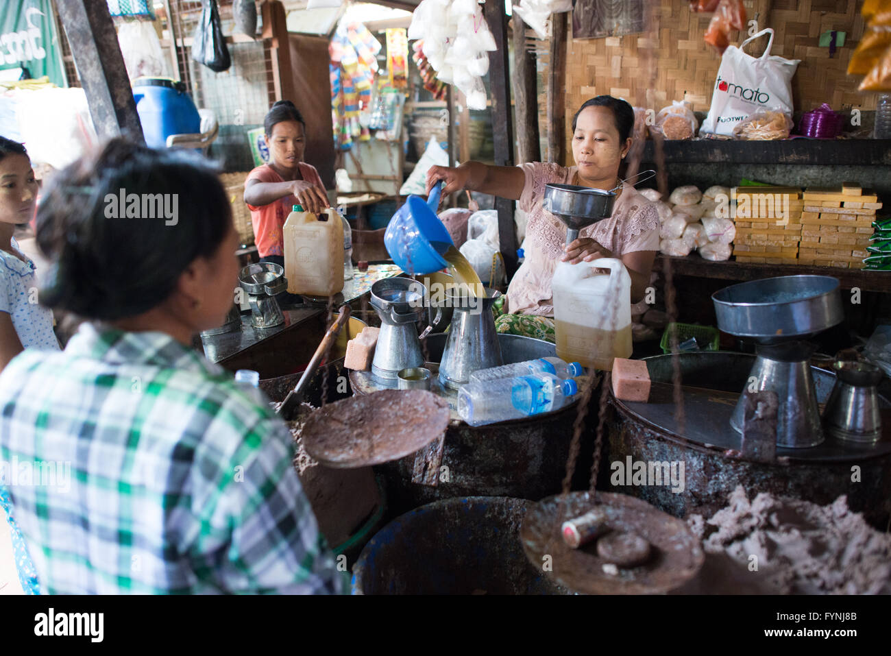 Venditore di olio da cucina che versa attraverso un imbuto metallico contenitori Nyaung U Myanmar | NYAUNG-U, Myanmar — Un venditore versa olio da cucina attraverso un imbuto metallico da un contenitore all’ingrosso al mercato di Nyaung-U. La città funge da centro commerciale per la zona arida centrale del Myanmar, dove arachidi resistenti alla siccità e sesamo vengono pressati per produrre oli non raffinati centrali per la cucina birmana. I clienti aspettano nelle vicinanze mentre scaffali di articoli per la casa delimitano la bancarella. Foto Stock