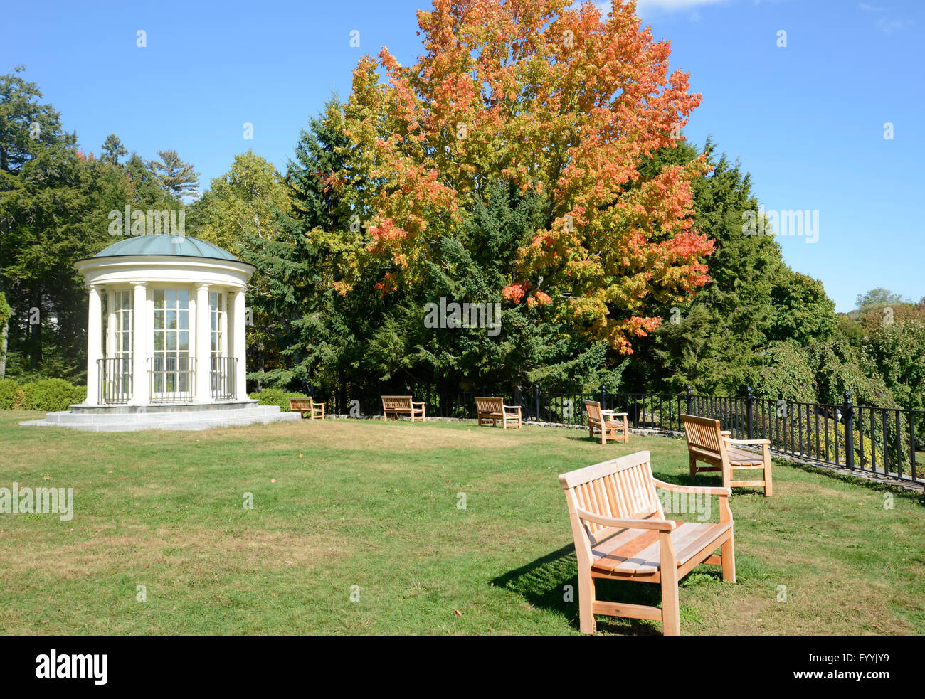 Round gazebo in legno e panchine da un nero recinzione in ferro. Vi è una zona di erba e alberi con foglie di iniziare a cambiare colore Foto Stock