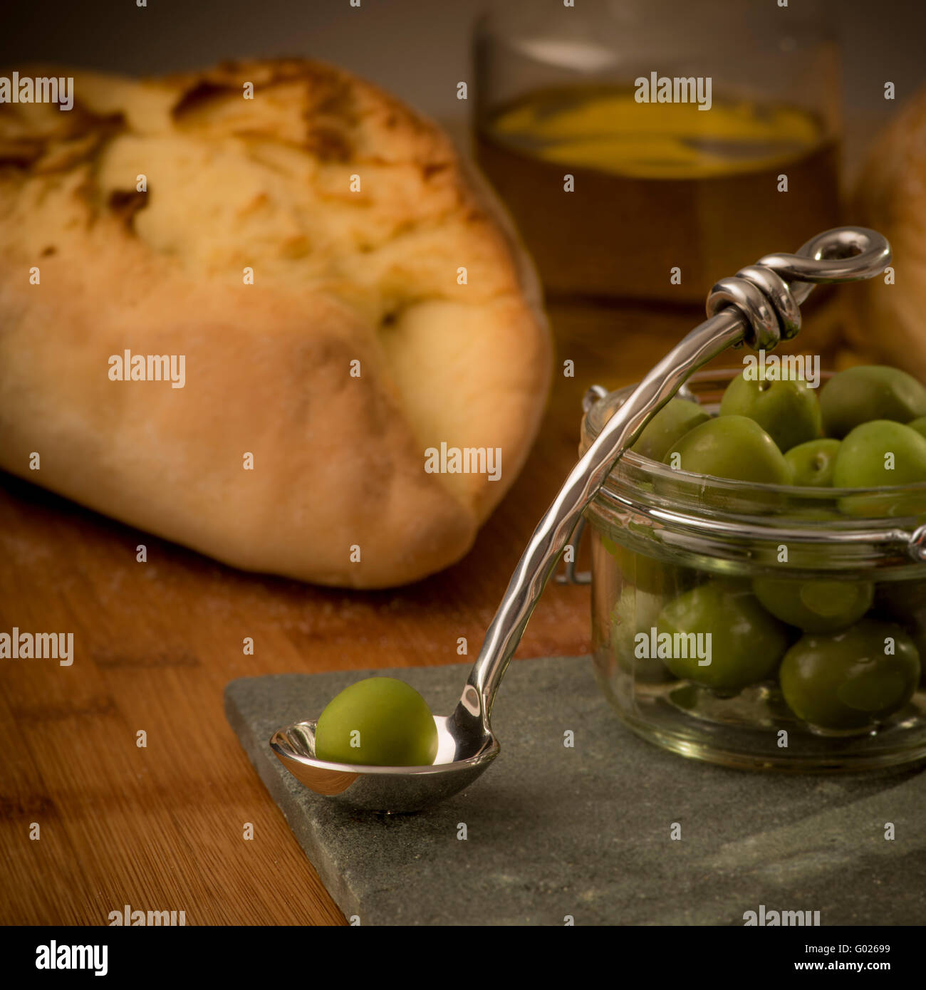 Vaso di olive verdi con un unico olio sul cucchiaio e olio di oliva con pane in background. Foto Stock