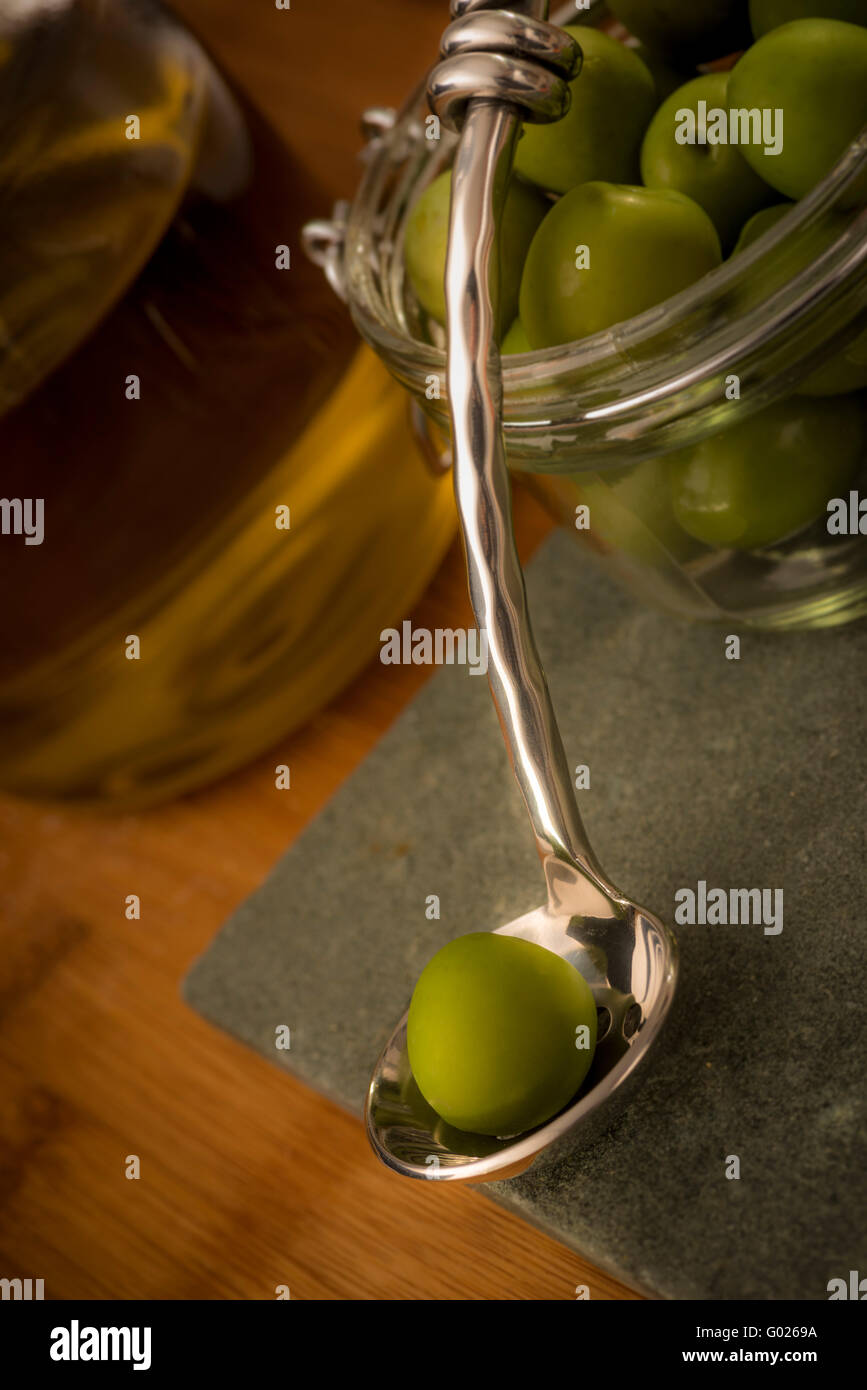 Vaso di olive verdi con un unico olio sul cucchiaio e olio d'oliva in background. Foto Stock