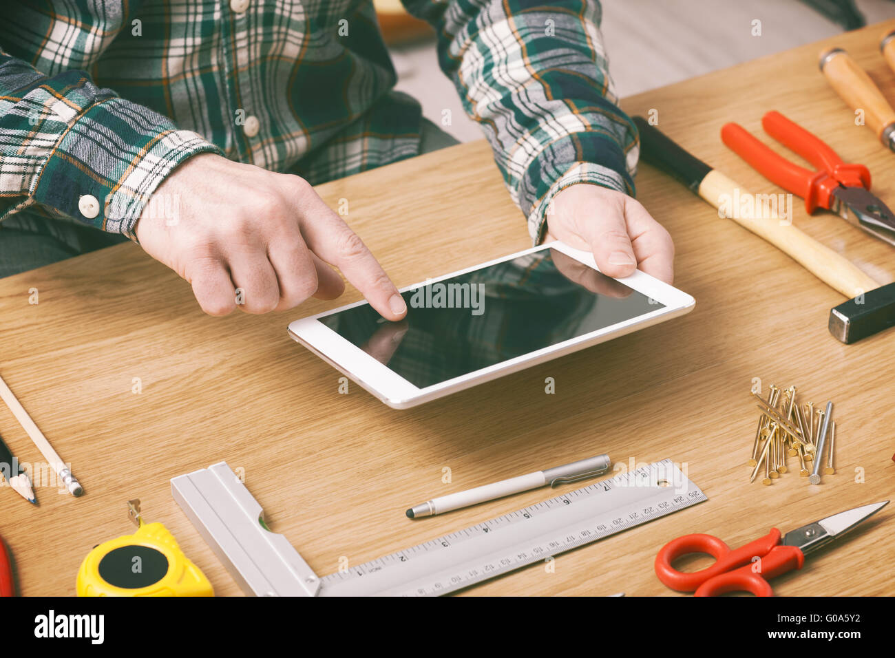 Uomo al lavoro su un progetto fai da te a casa con una tavoletta digitale, falegnameria e di strumenti di costruzione su di un tavolo di lavoro, mani vicino fino Foto Stock