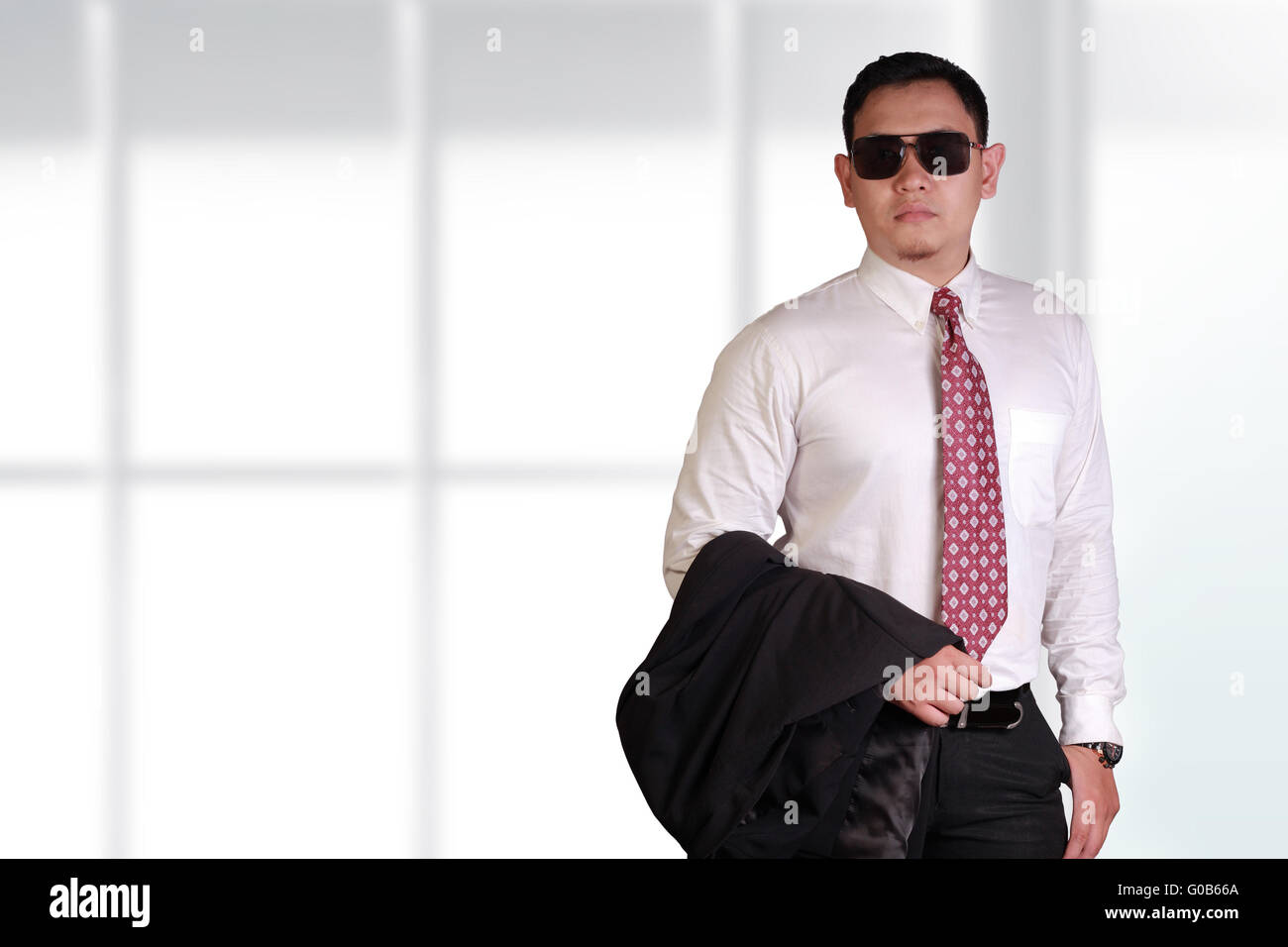 Giovane e orgoglioso uomo d'affari asiatici indossando occhiali nero tenendo la sua giacca tuta sul suo braccio su sfondo lucido Foto Stock