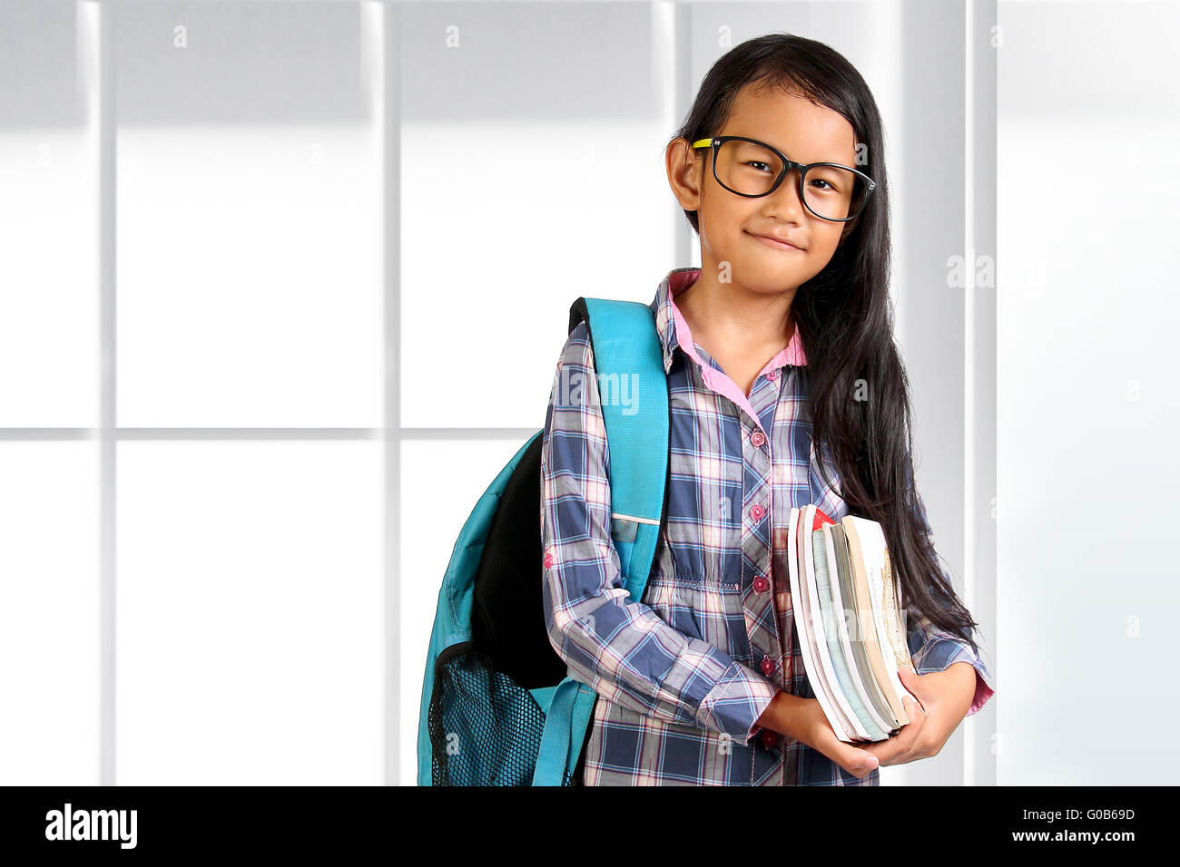 Giovani studenti asiatici ragazza con gli occhiali e zaino in piedi e sorridente mentre si tiene libri su sfondo lucido Foto Stock