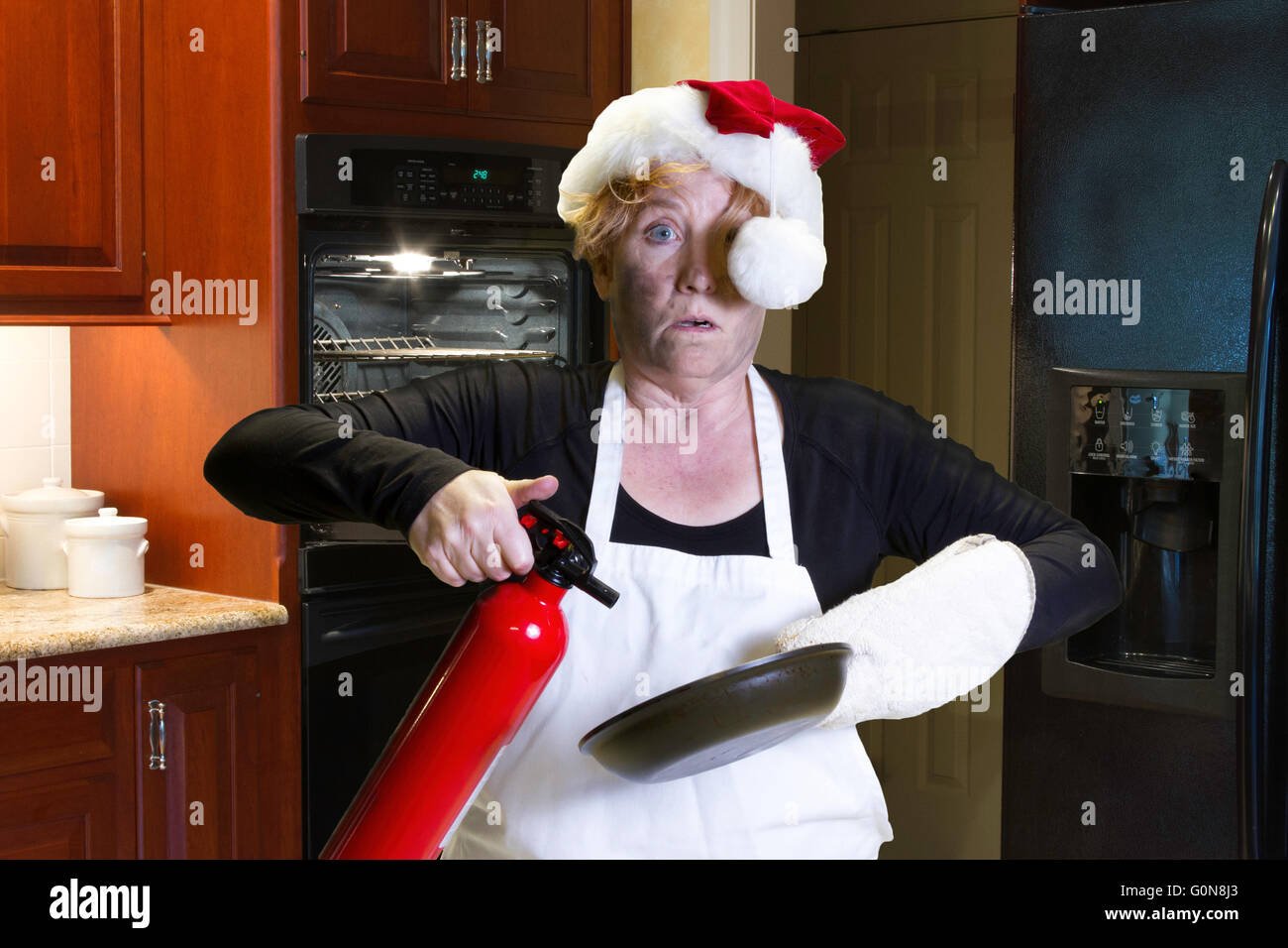 Donna matura con firmato faccia tenendo pan e estintore con santa hat e grembiule in piedi in cucina. Foto Stock