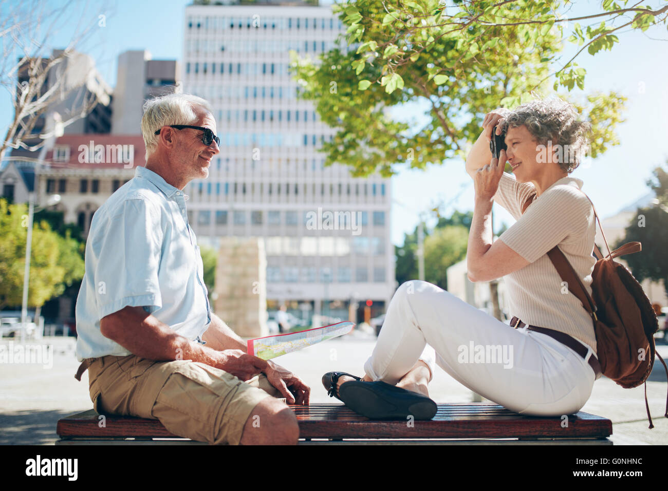 Vista laterale del senior uomo seduto all'aperto su un banco e la donna prendendo le sue foto con la fotocamera digitale. Coppia senior divertendosi Foto Stock