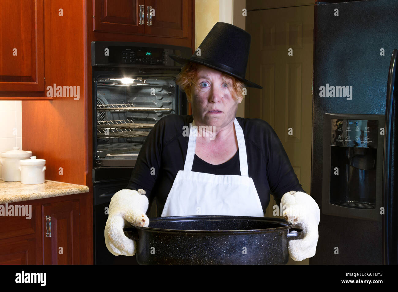 Donna in piedi in cucina indossando pellegrino hat e grembiule con bruciature viso e capelli tenendo una teglia. Foto Stock