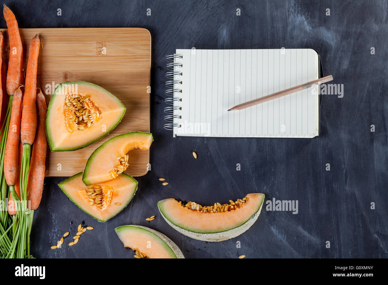 Fette di melone e mazzo di carote sul bordo di taglio sulla lavagna nera con i notebook Foto Stock