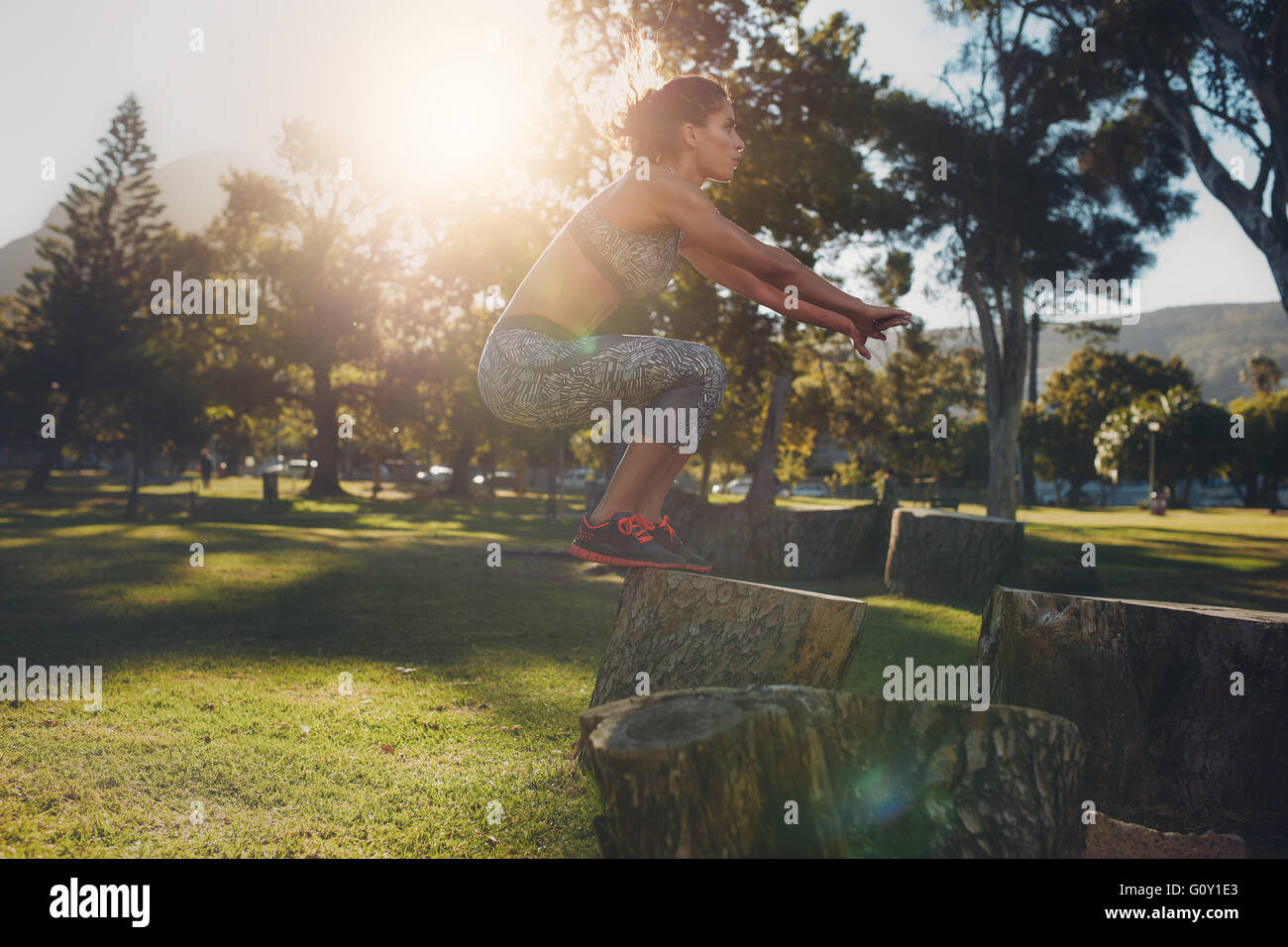 Colpo di una giovane donna di saltare su log in legno presso il parco. Sportive fare esercizio in natura in una giornata di sole. La casella salta. Foto Stock