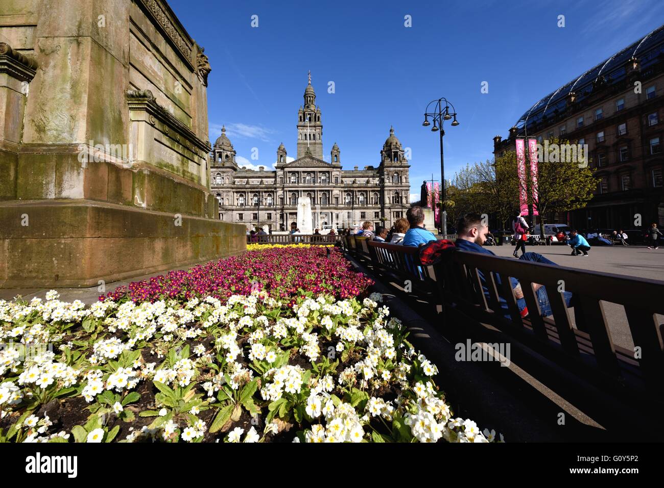 Persone sedersi in George Square e crogiolatevi al sole primaverile, con Glasgow City Chambers in background. Foto Stock