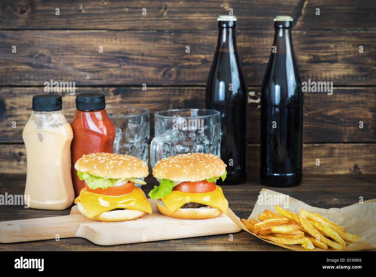 Due cheeseburger con due bottiglie di birra scura e due tazze a vuoto su sfondo di legno Foto Stock