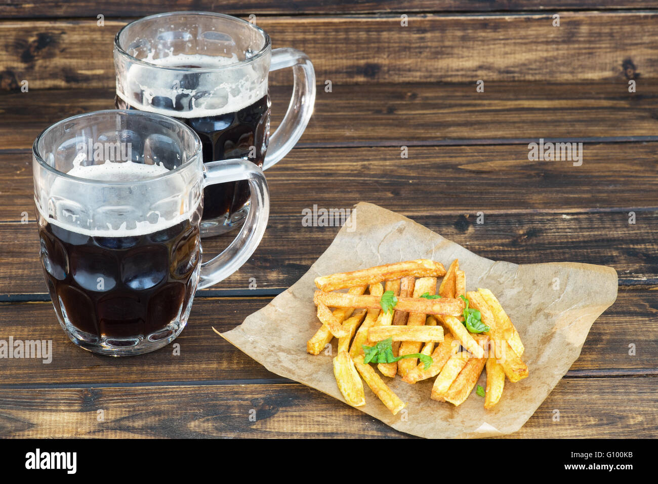 Due boccali di birra scura con patate fritte su sfondo di legno Foto Stock