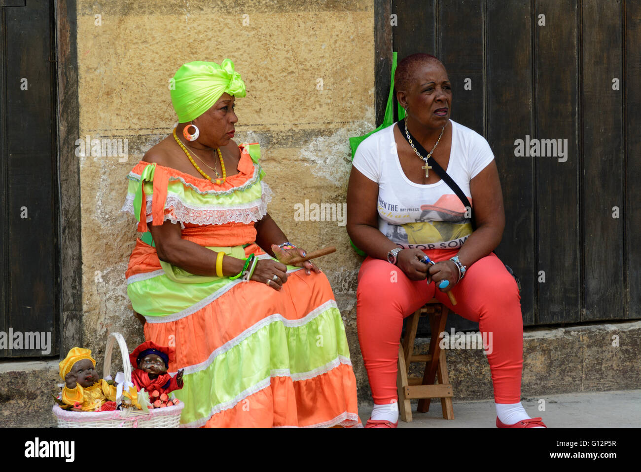 Un cubano doll venditore indossando il costume tradizionale e fumare un sigaro si rilassa con un compagno in Avana Vecchia, Havana, Cuba Foto Stock