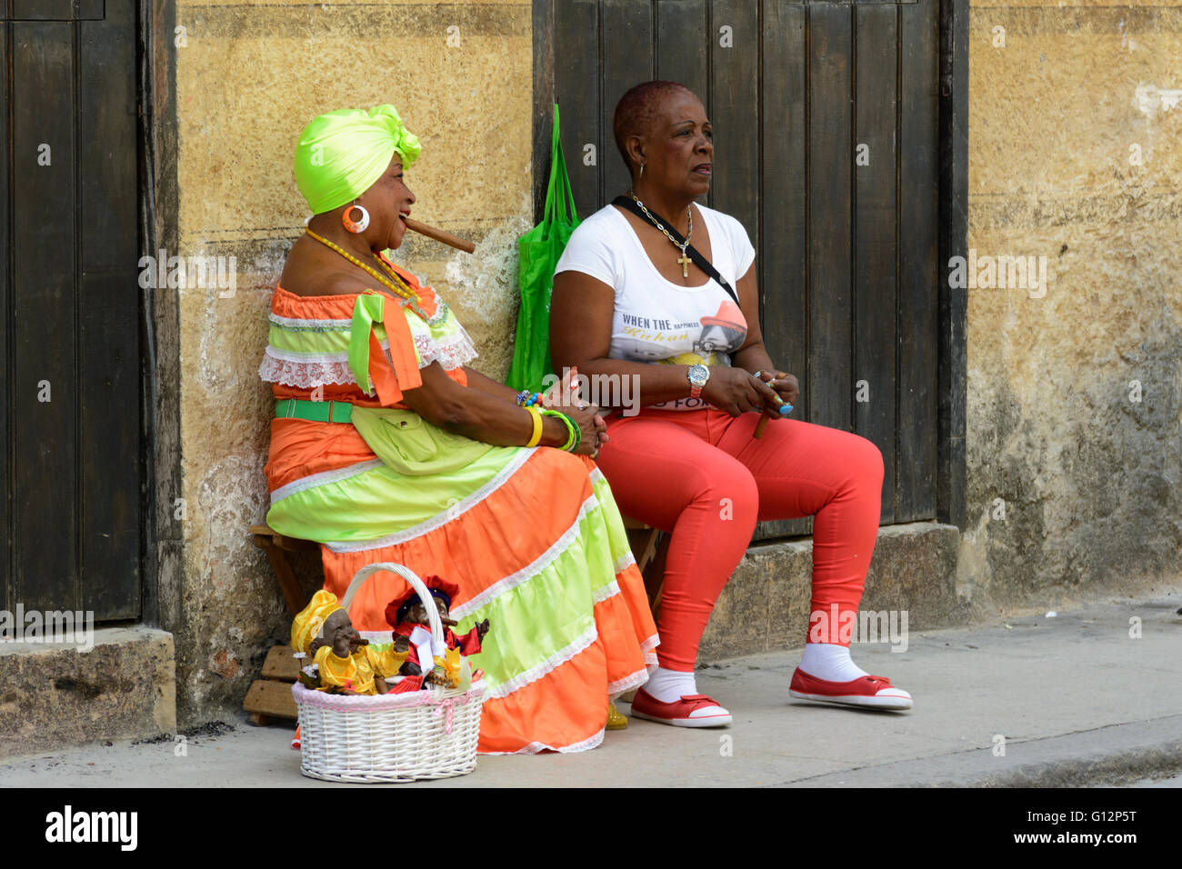 Un cubano doll venditore indossando il costume tradizionale e fumare un sigaro si rilassa con un compagno in Avana Vecchia, Havana, Cuba Foto Stock