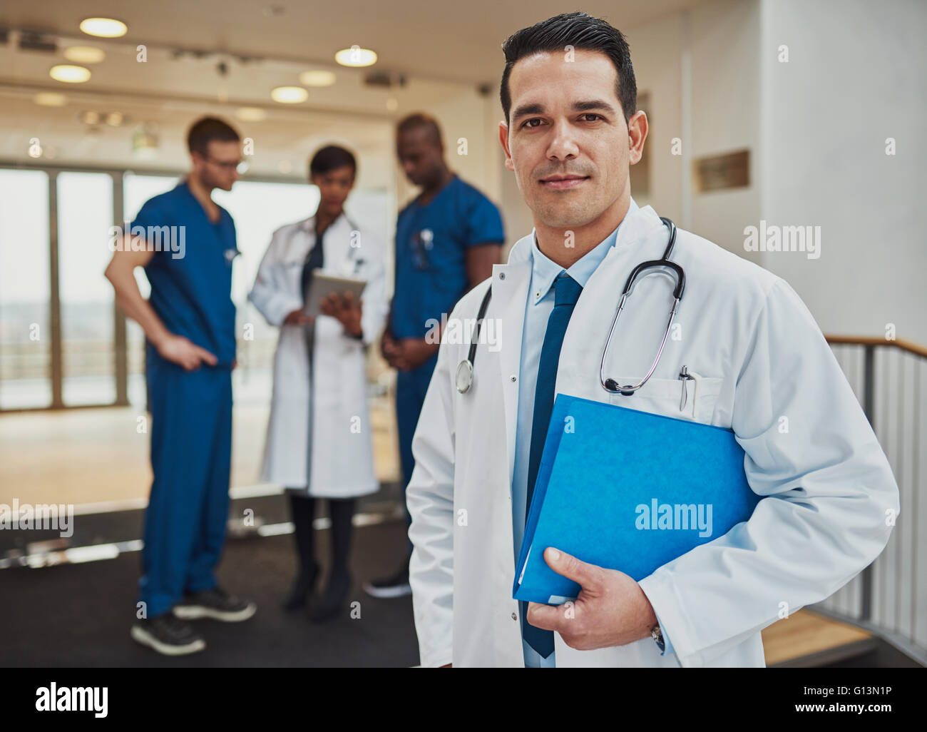 Considerato i giovani ispanici medico in ospedale in piedi guardando la telecamera con un file sotto il suo braccio come la sua squadra multirazziale Foto Stock