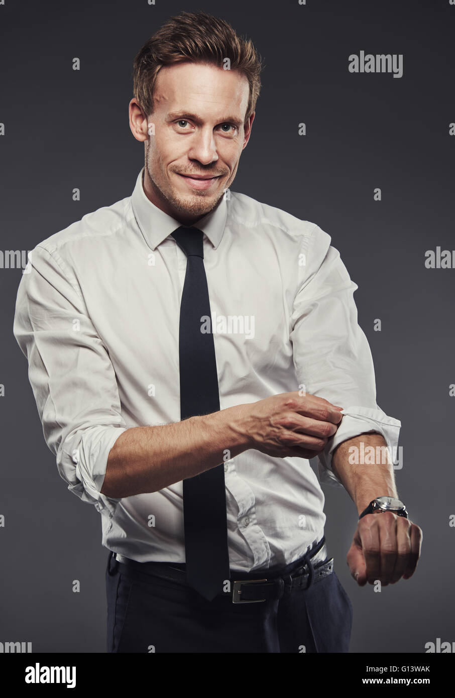 Bello imprenditore arrotolare la sua maglietta maniche mentre guardando la telecamera con un sorriso amichevole e sfondo grigio Foto Stock