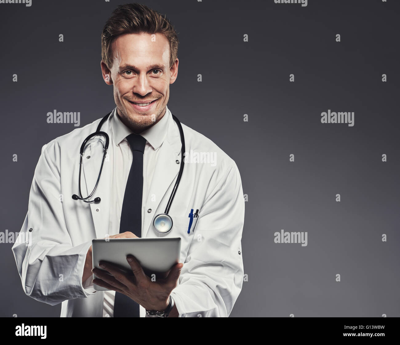 Sorridente medico con stetoscopio e compressa bianca da indossare camice e black tie si erge contro uno sfondo scuro Foto Stock