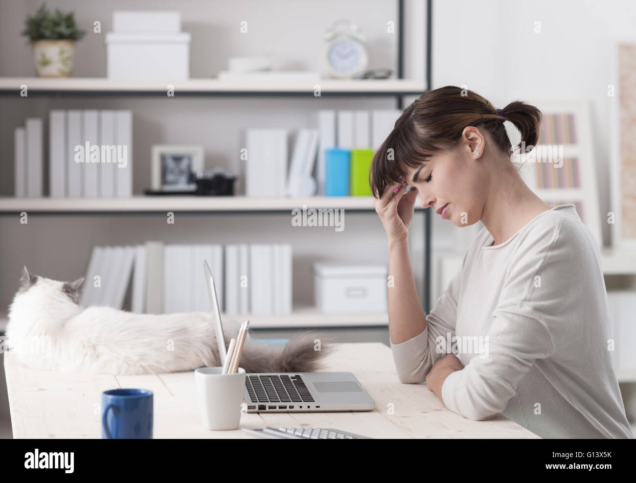 La donna a casa, seduti alla scrivania e lavorare con un computer portatile ha un mal di testa e toccando la fronte, un gatto sdraiato su Foto Stock