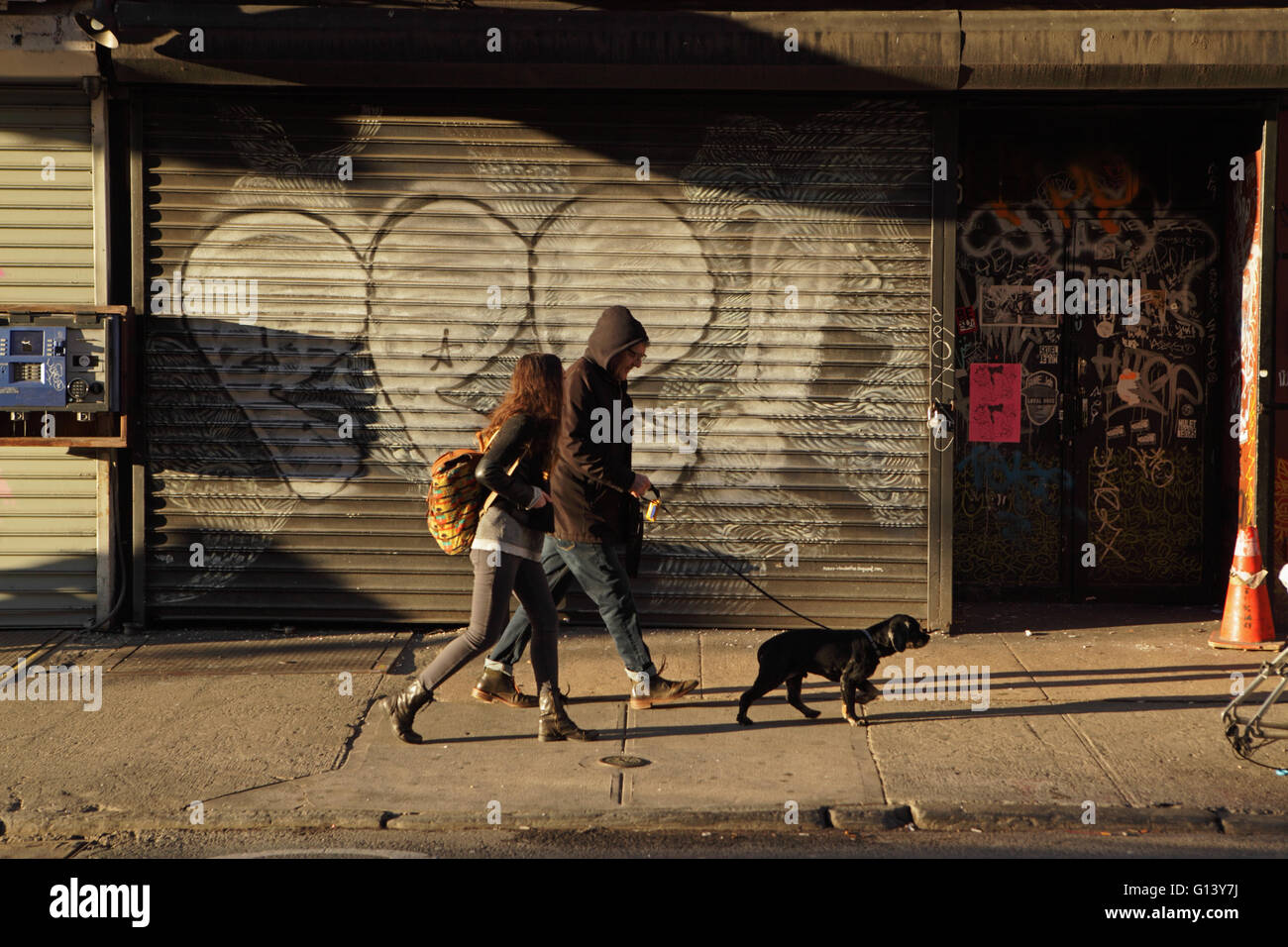 L'uomo con il cane al guinzaglio e una ragazza a piedi verso est lungo Rivington Street sulla Lower East Side di New York come il sole che sorge Foto Stock