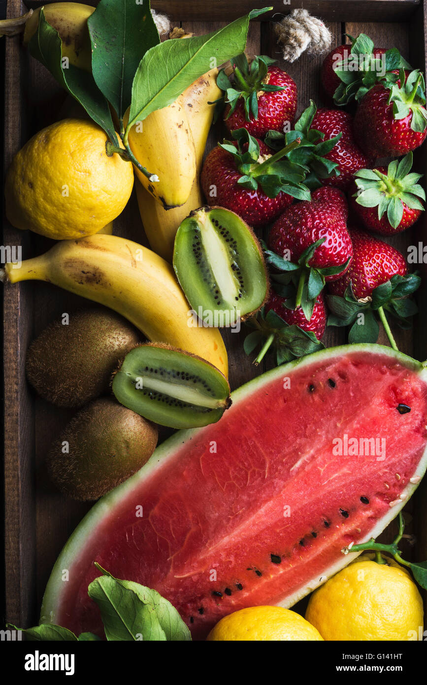 Varie colorata frutta tropicale selezione nel vassoio di legno su sfondo scuro, vista dall'alto. Anguria, fragola, limoni e kiwi Foto Stock