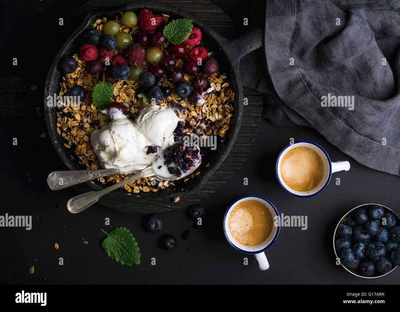 Una sana prima colazione. Oat granola sgretolarsi con frutti di bosco freschi, semi e gelati in padella di ferro padella in legno scuro e scheda di tazze Foto Stock