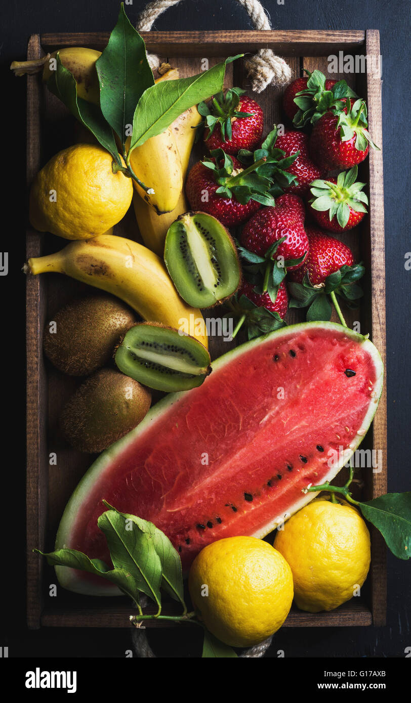 Varie colorata frutta tropicale selezione nel vassoio di legno su sfondo scuro, vista dall'alto. Anguria, fragola, limoni e kiwi Foto Stock