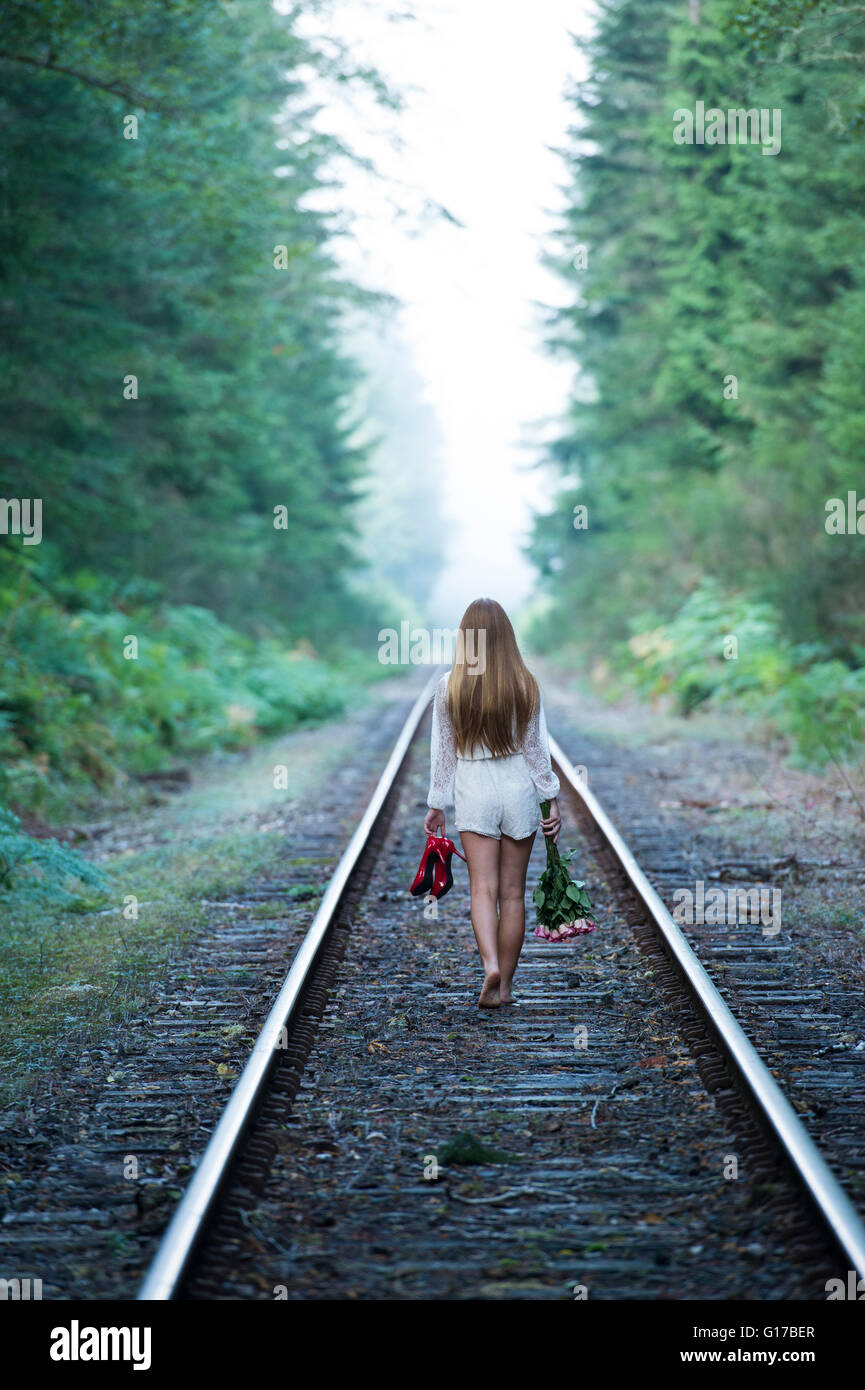 Ragazza adolescente, passeggiate lungo via treno, tenendo red shoes e fiori, vista posteriore Foto Stock