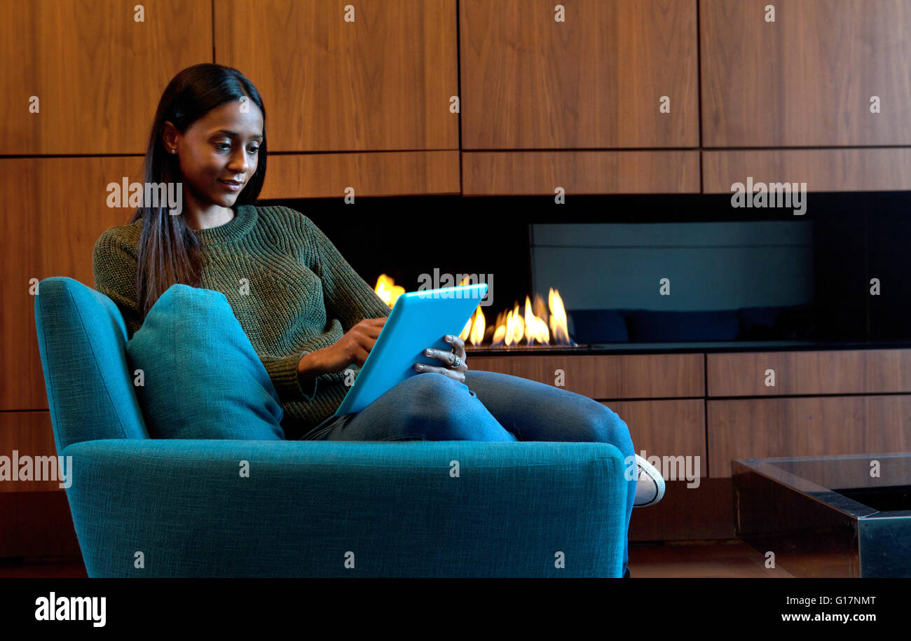 Donna in poltrona nella parte anteriore del cabinet in legno e fuoco aperto, utilizzando tavoletta digitale Foto Stock