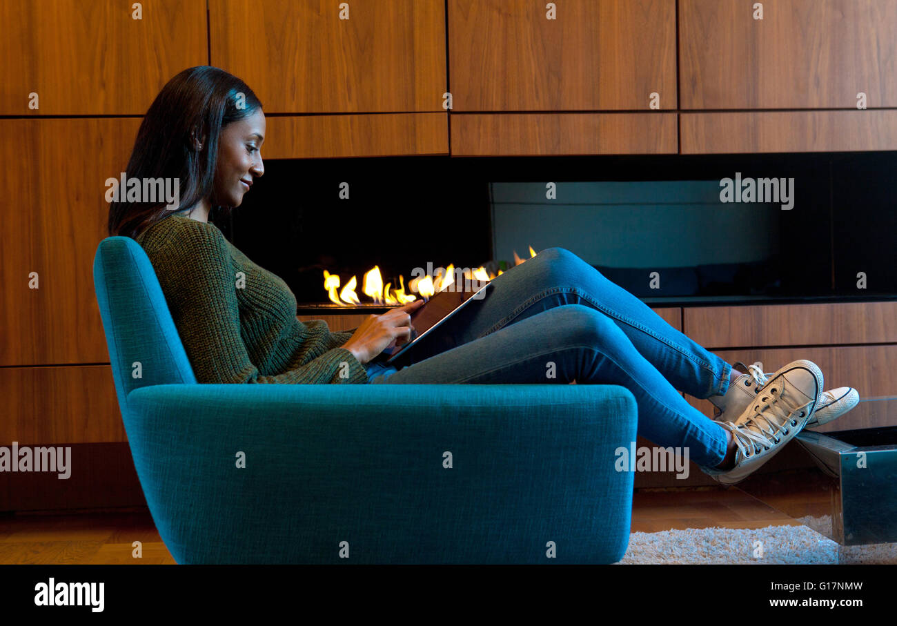 Donna in poltrona nella parte anteriore del cabinet in legno e fuoco aperto, utilizzando tavoletta digitale Foto Stock