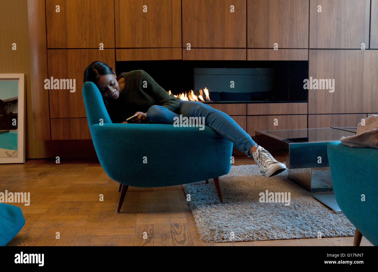 Donna in poltrona nella parte anteriore del cabinet in legno e fuoco aperto utilizza lo smartphone Foto Stock