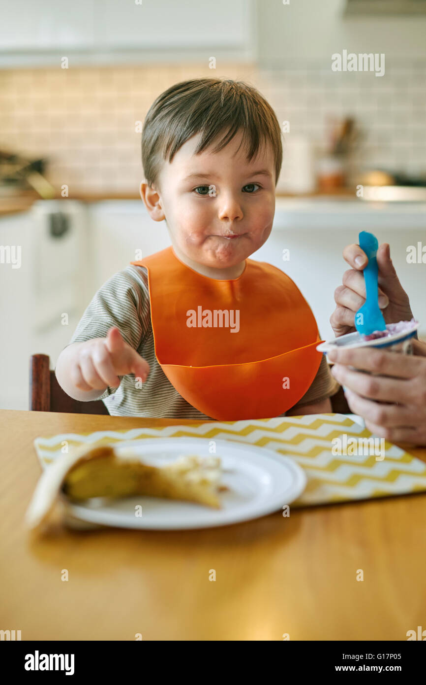 Baby boy seduta a tavola essendo alimentato yogurt da madre Foto Stock