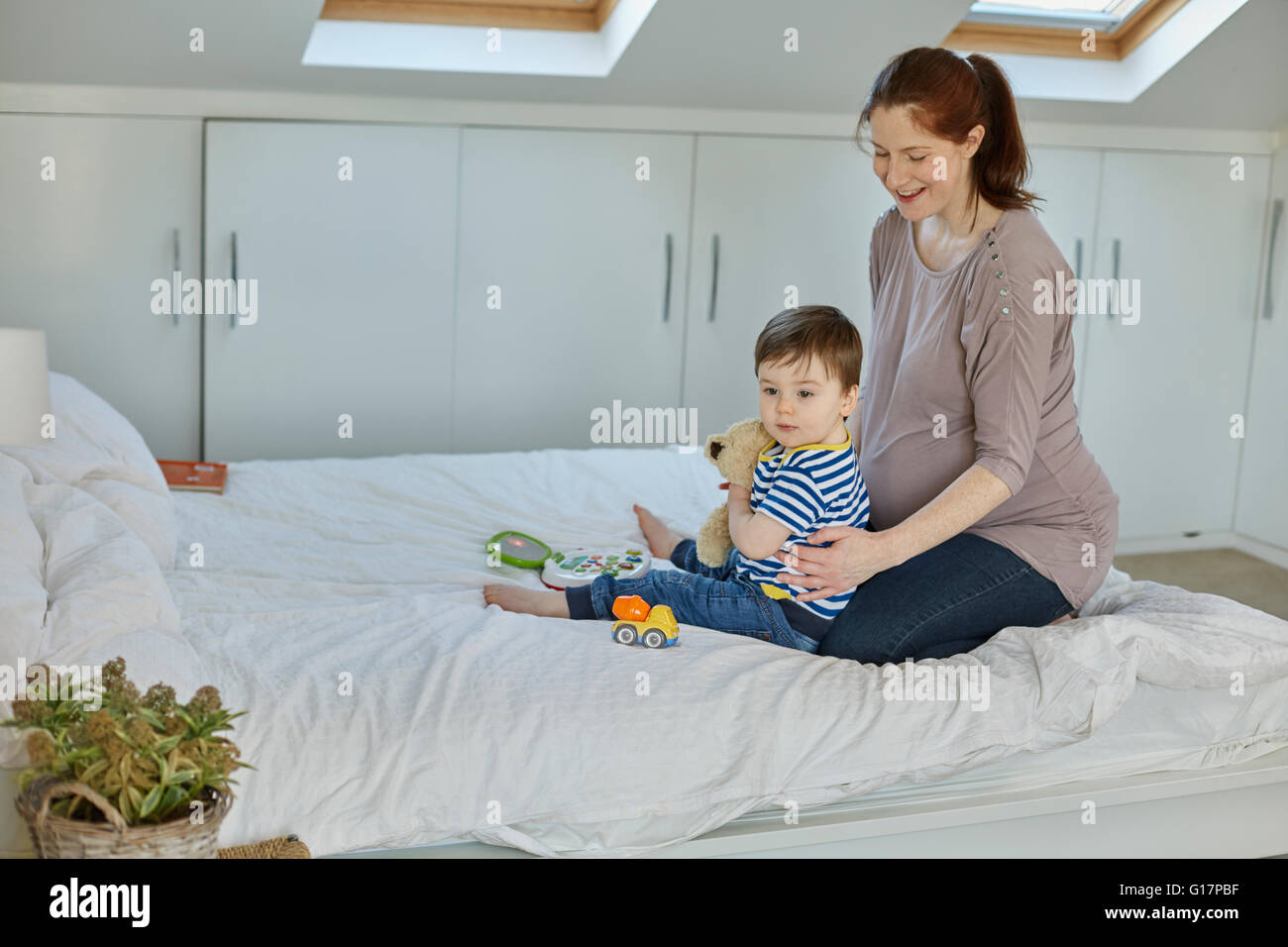 Donna incinta seduta sul letto con baby boy cuddling orsacchiotto Foto Stock