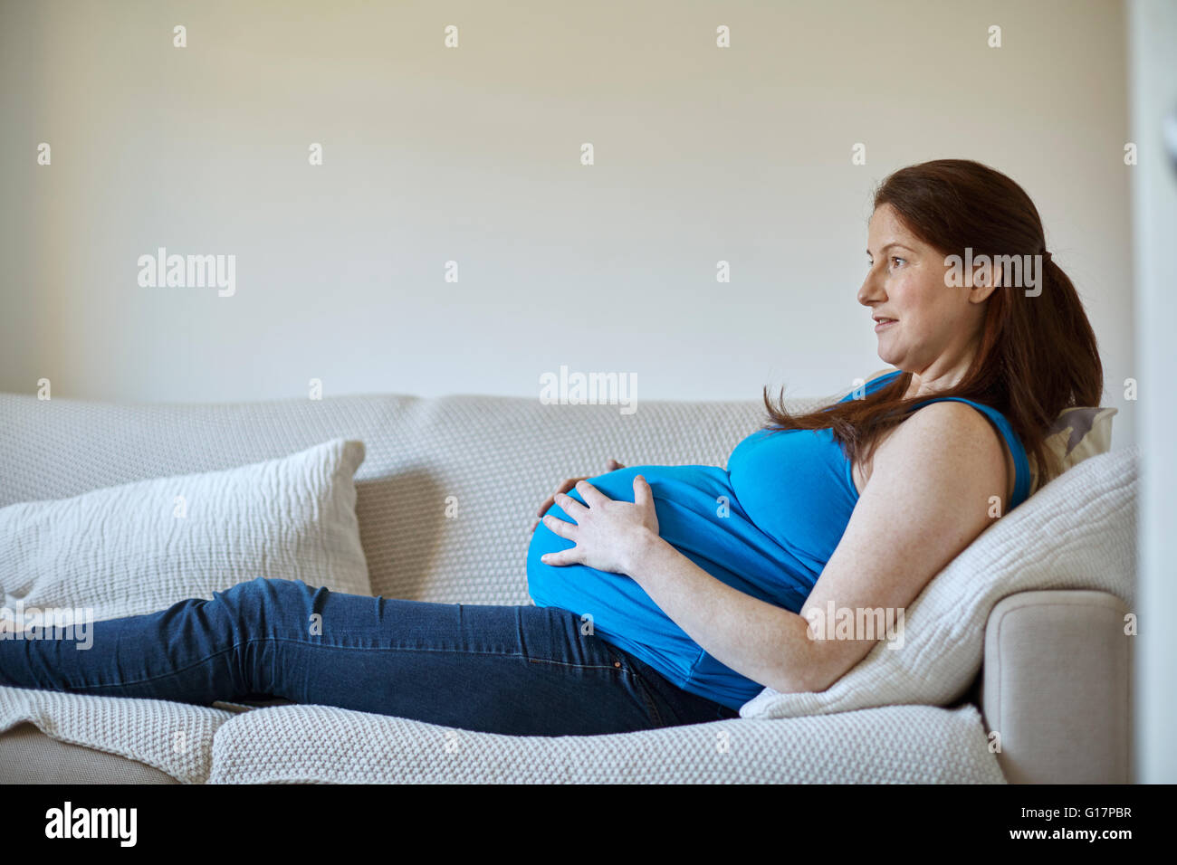 Vista laterale della donna incinta holding stomaco, rilassante sul divano guardando lontano sorridente Foto Stock