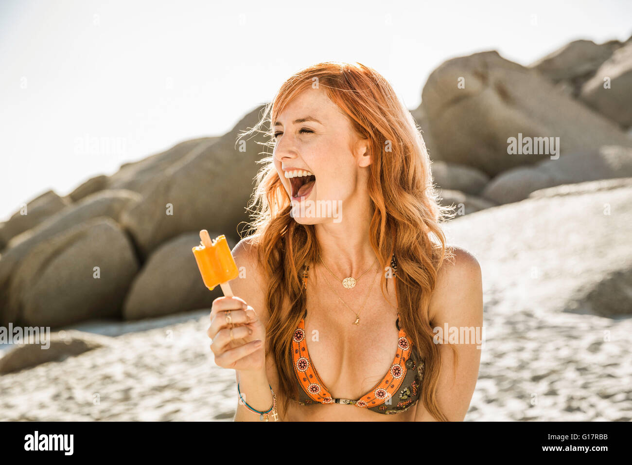 Ridendo donna con lunghi capelli rossi di mangiare per lecca-lecca di ghiaccio sulla spiaggia, Cape Town, Sud Africa Foto Stock