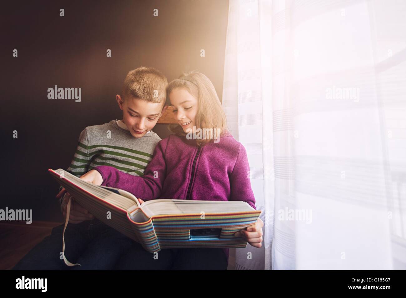 Un ragazzo e una ragazza gemelli guardando album di foto accanto alla finestra Foto Stock
