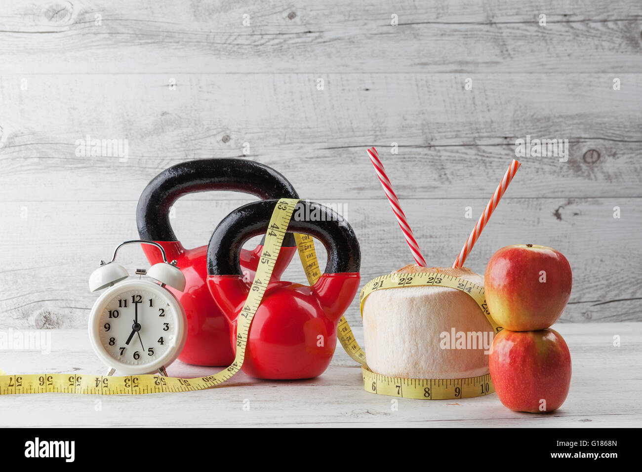 Due kettlebells rosso con nastro di misurazione, bere noce di cocco, cannucce, mele e vintage orologio sul bianco rustico tavolo in legno. Healt Foto Stock