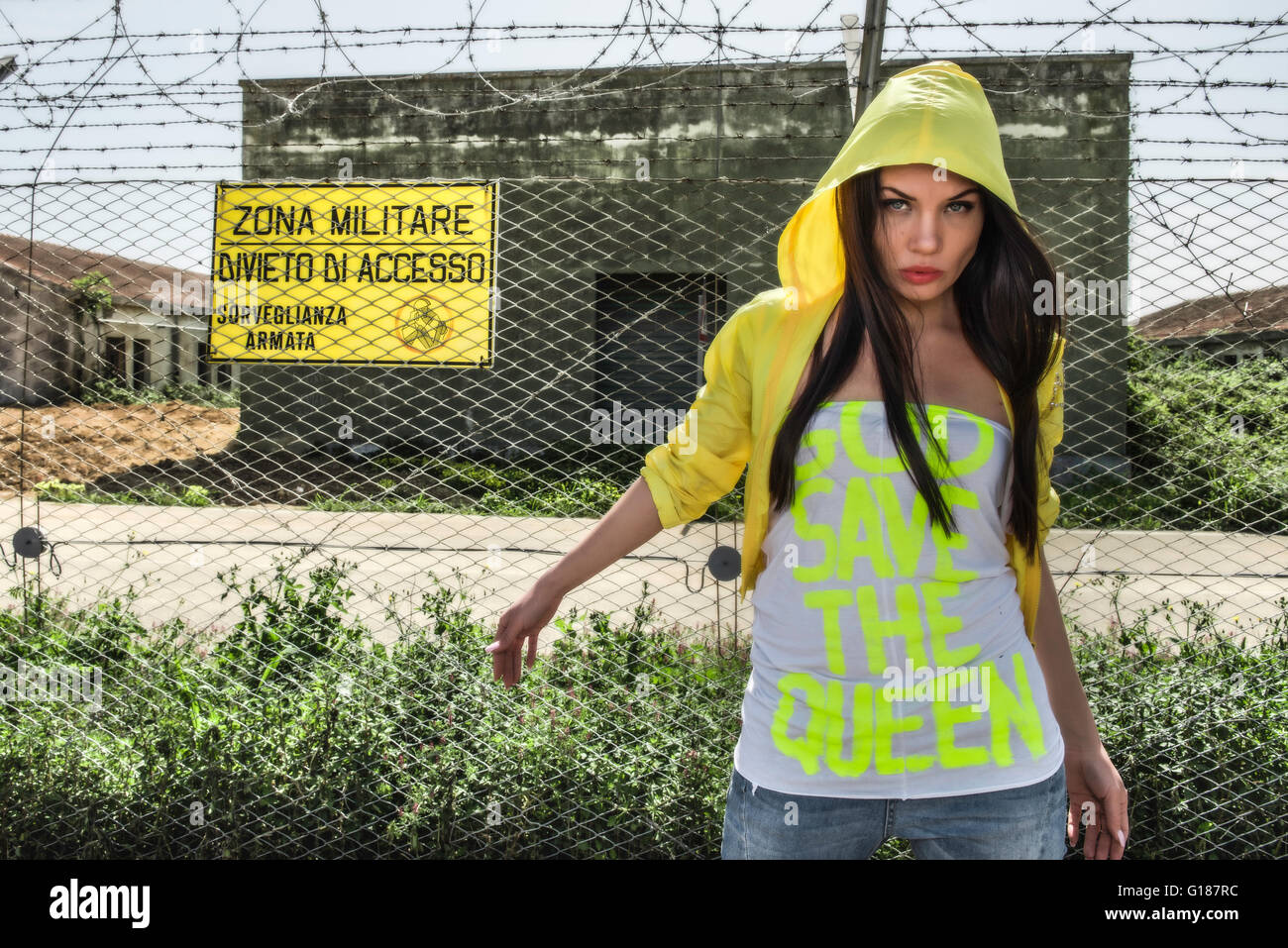 Modello indossa una giacca impermeabile giallo e una camicia la lettura Dio salvi la regina, in piedi al di fuori di un composto militare Foto Stock