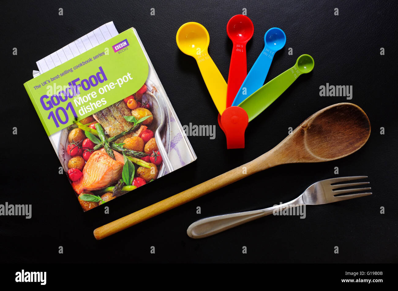 Un piatto scena laici di un libro di cucina, misurini, un cucchiaio di legno e una forca impostato su sfondo nero. Foto Stock
