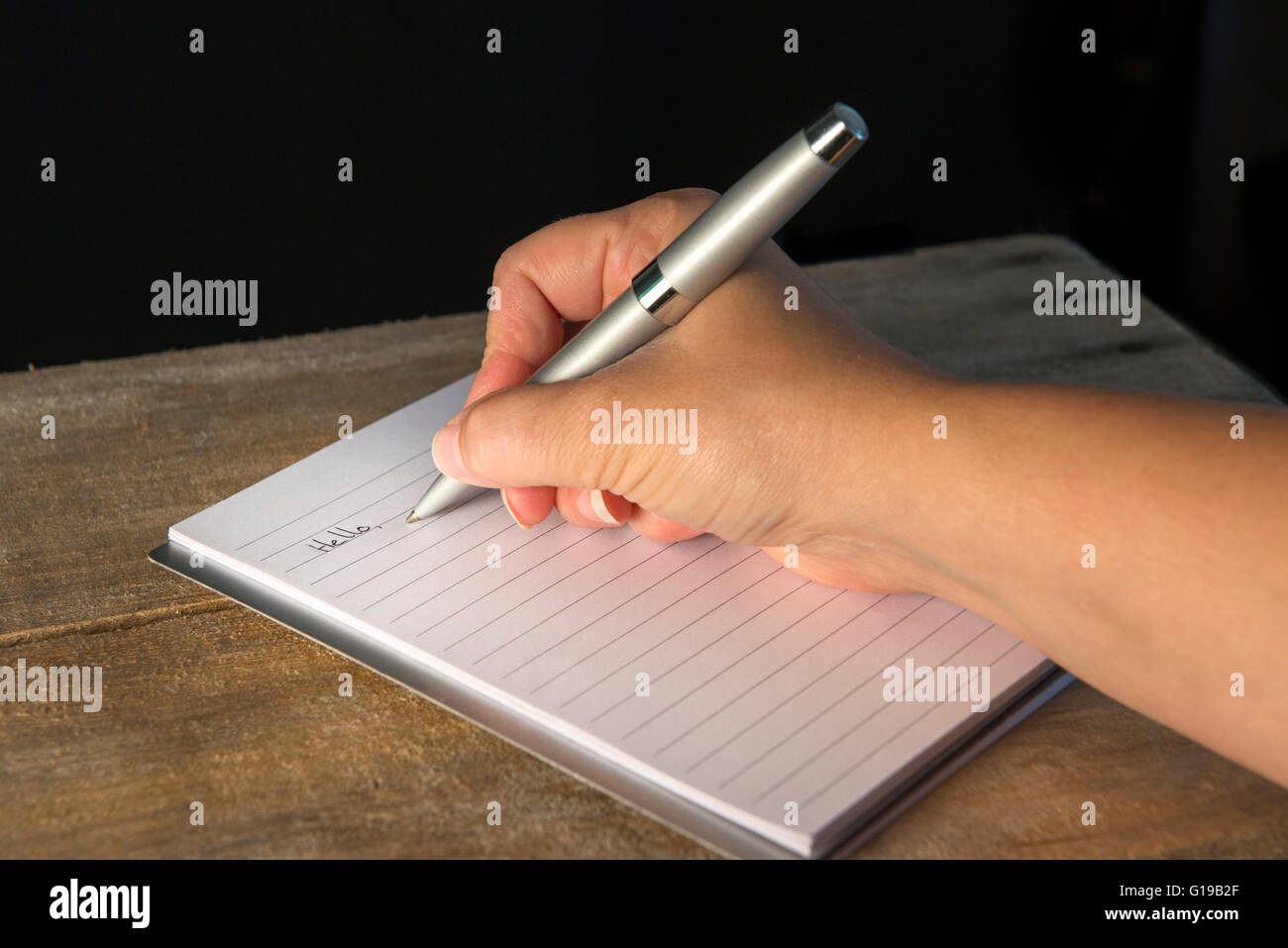 Primo piano di una scrittura a mano con una penna argentata su un blocco note foderato, su una superficie in legno con sfondo scuro, concetto di scrittura, inserimento nel Journal. Foto Stock