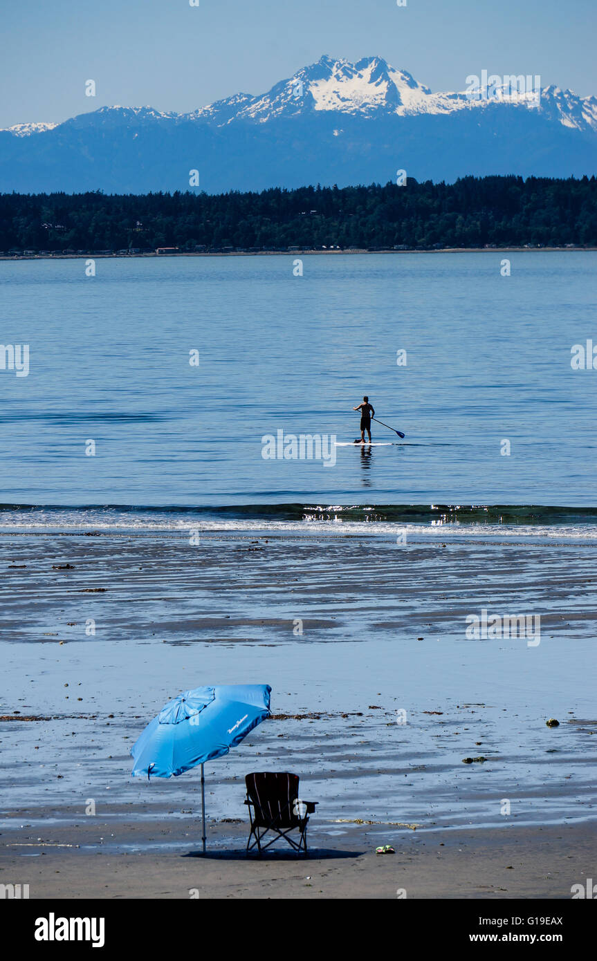 Uomo sulla paletta passa scheda di sedie e ombrellone blu sulla sabbia a bassa marea sul Puget Sound con la snowy Olympic Mountain Range e Foto Stock