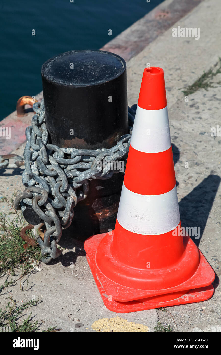 Rosso di avvertimento a strisce cono stradale si erge nei pressi di bollard ormeggio nel porto di mare Foto Stock